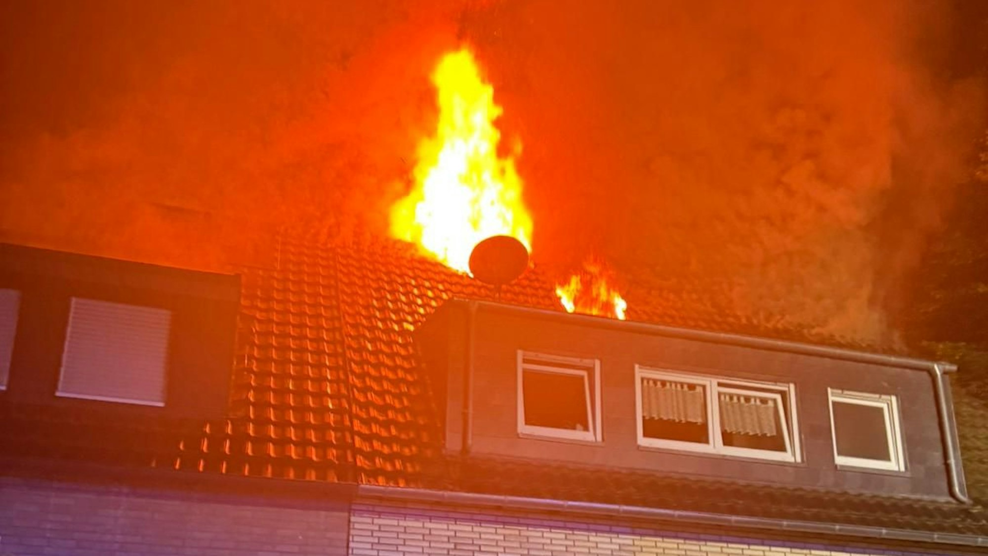 Das Foto zeigt den Dachstuhl mit Gaube eines Mehrfamilienhauses. Aus dem Dach schlagen hohe Flammen.