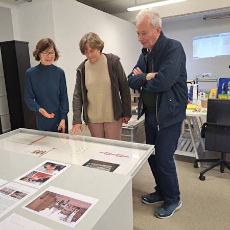 Zwei Frauen und ein Mann schauen sich Kunstwerke, die vor ihnen liegen, an.