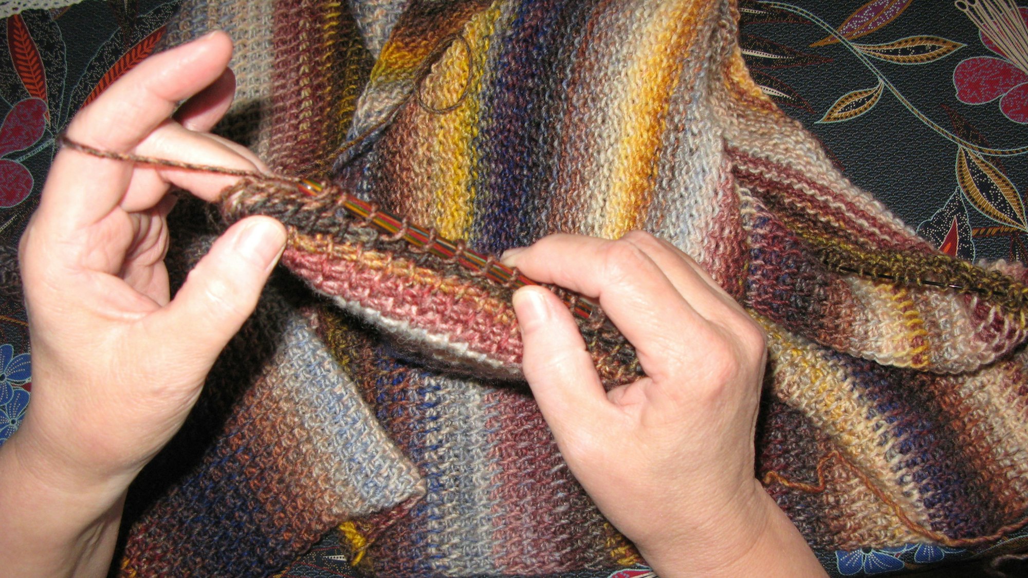 Zwei Hände halten einen Wollfaden und eine Häkelnadel, darunter liegt das bereits fertig gehäkelte Tuch.