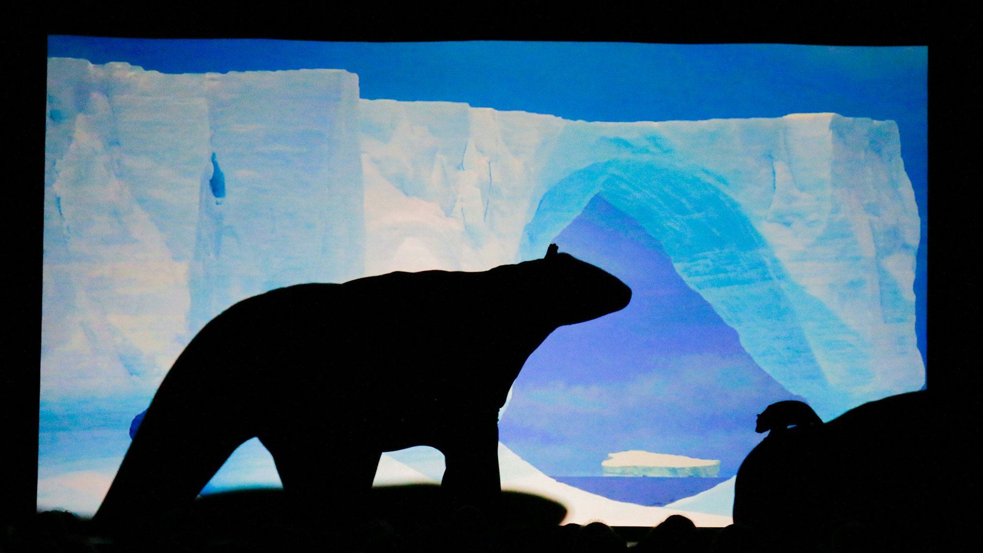 Auf einer Leinwand ist vor einer gemalten Polarlandschaft die Silhouette eines Eisbären zu sehen.