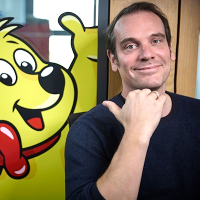 Konrad Lohrmann, Zeichner des Haribo-Goldbären, zeigt mit dem rechten Daumen hinter sich auf einen Haribo-Goldbär an einer Tür