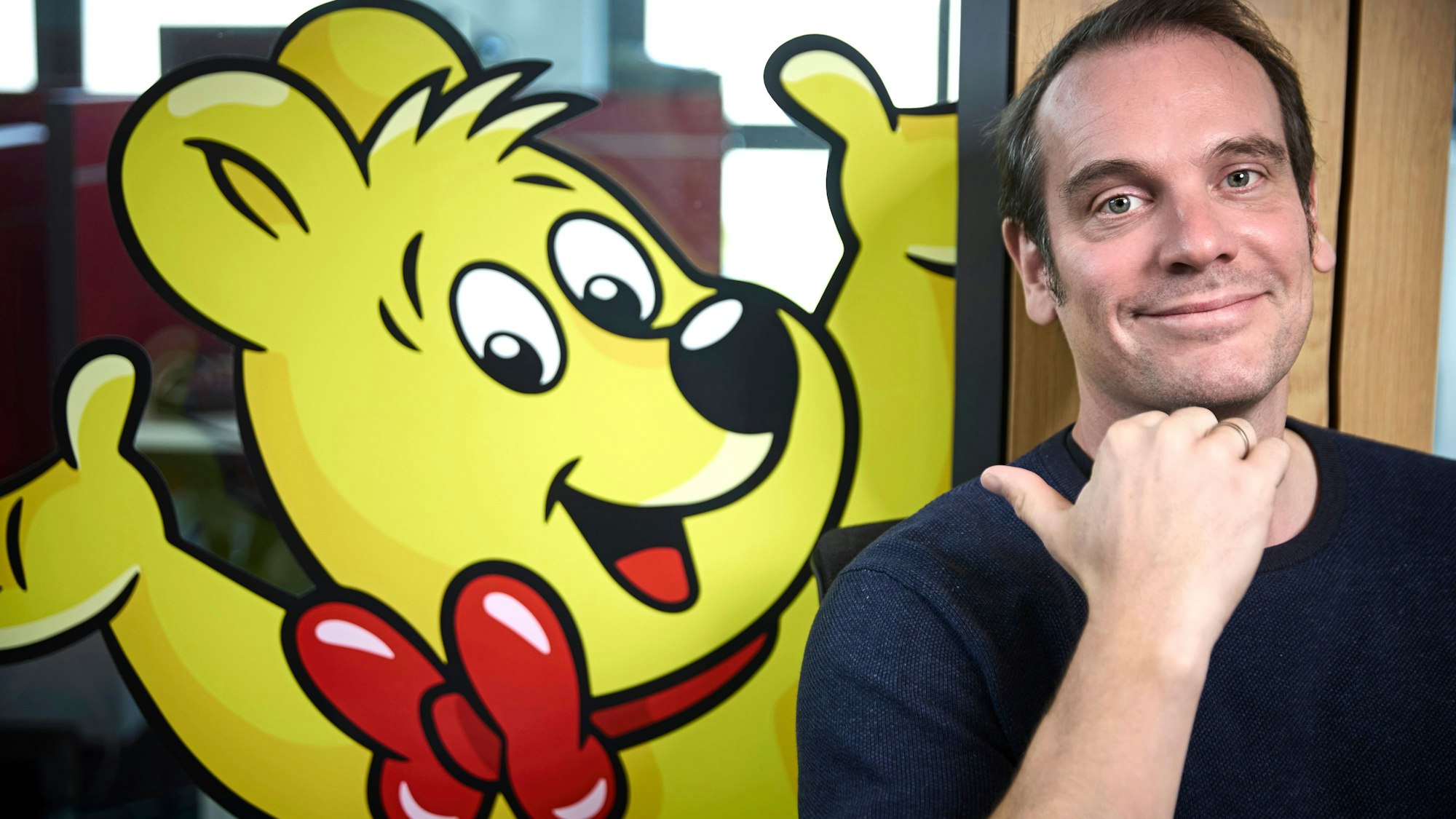 Konrad Lohrmann, Zeichner des Haribo-Goldbären, zeigt mit dem rechten Daumen hinter sich auf einen Haribo-Goldbär an einer Tür