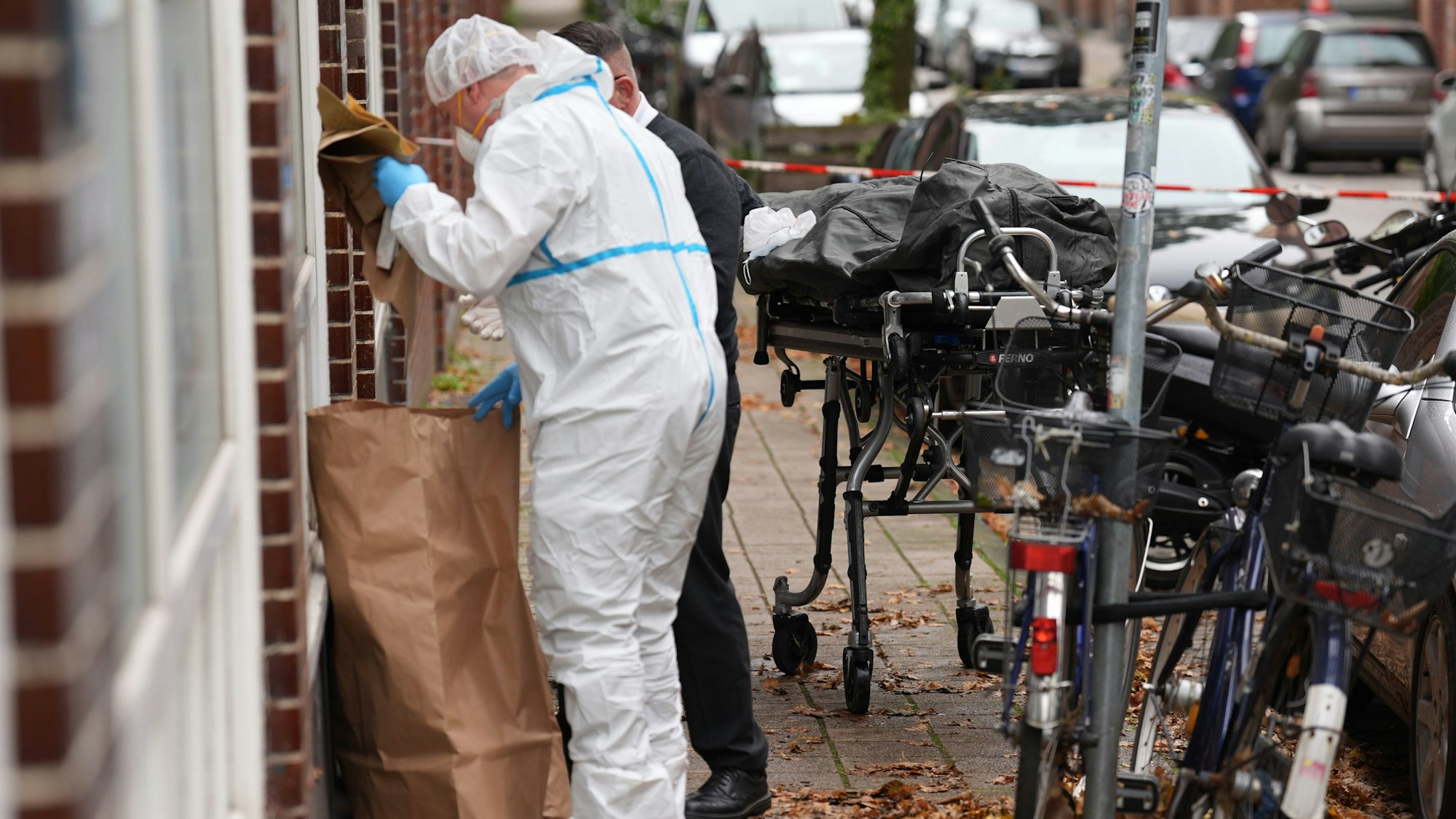 Ein Toter liegt in einem Leichensack (r) auf einer Trage neben einem Mitarbeiter der Kriminaltechnik und einem Bestatter vor einem Mehrfamilienhaus.