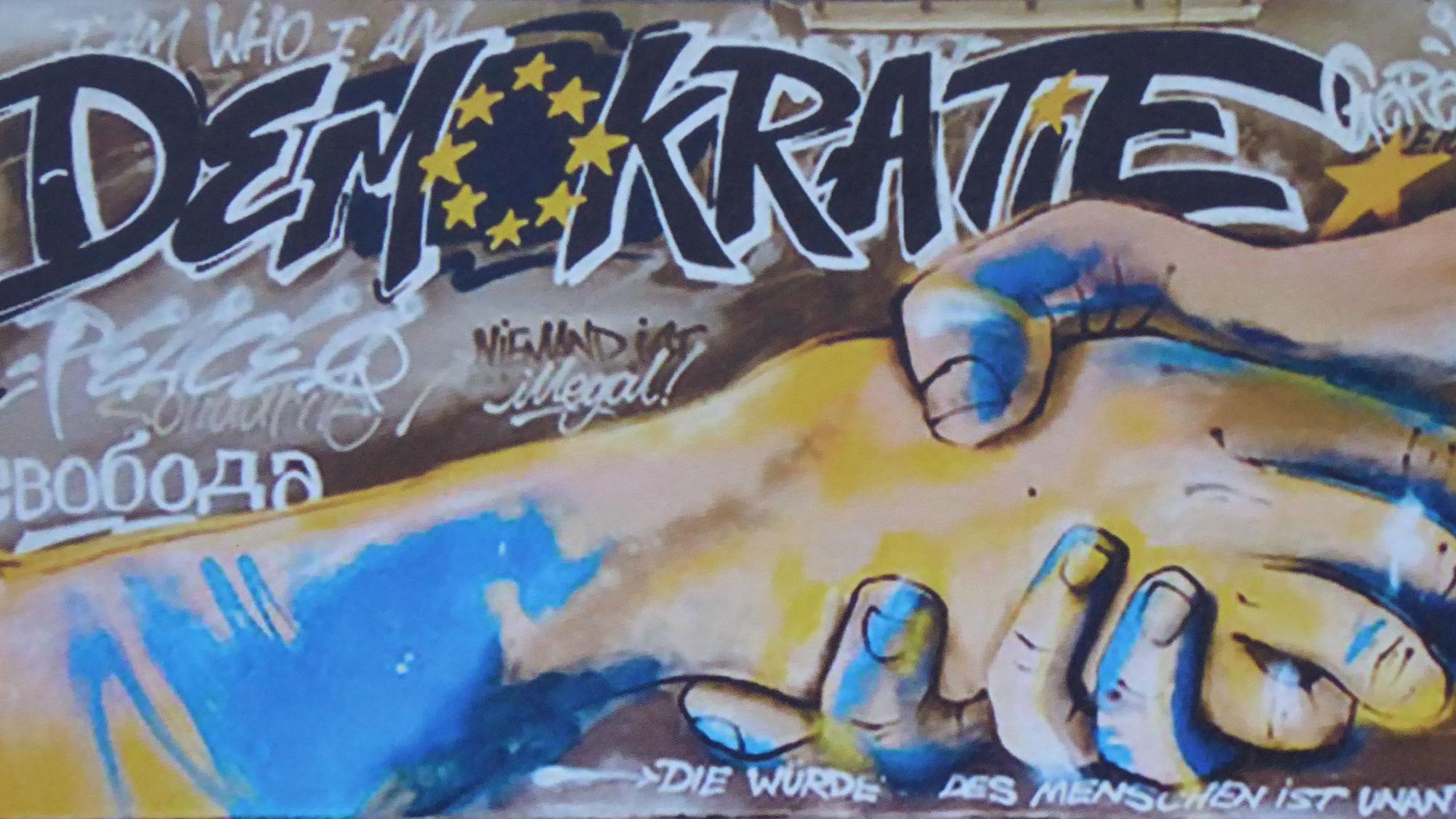 Ein Graffito mit einem Handschlag von zwei Händen und der Überschrift Demokratie mit EU-Sternenkreis an Stelle des Buchstaben o.