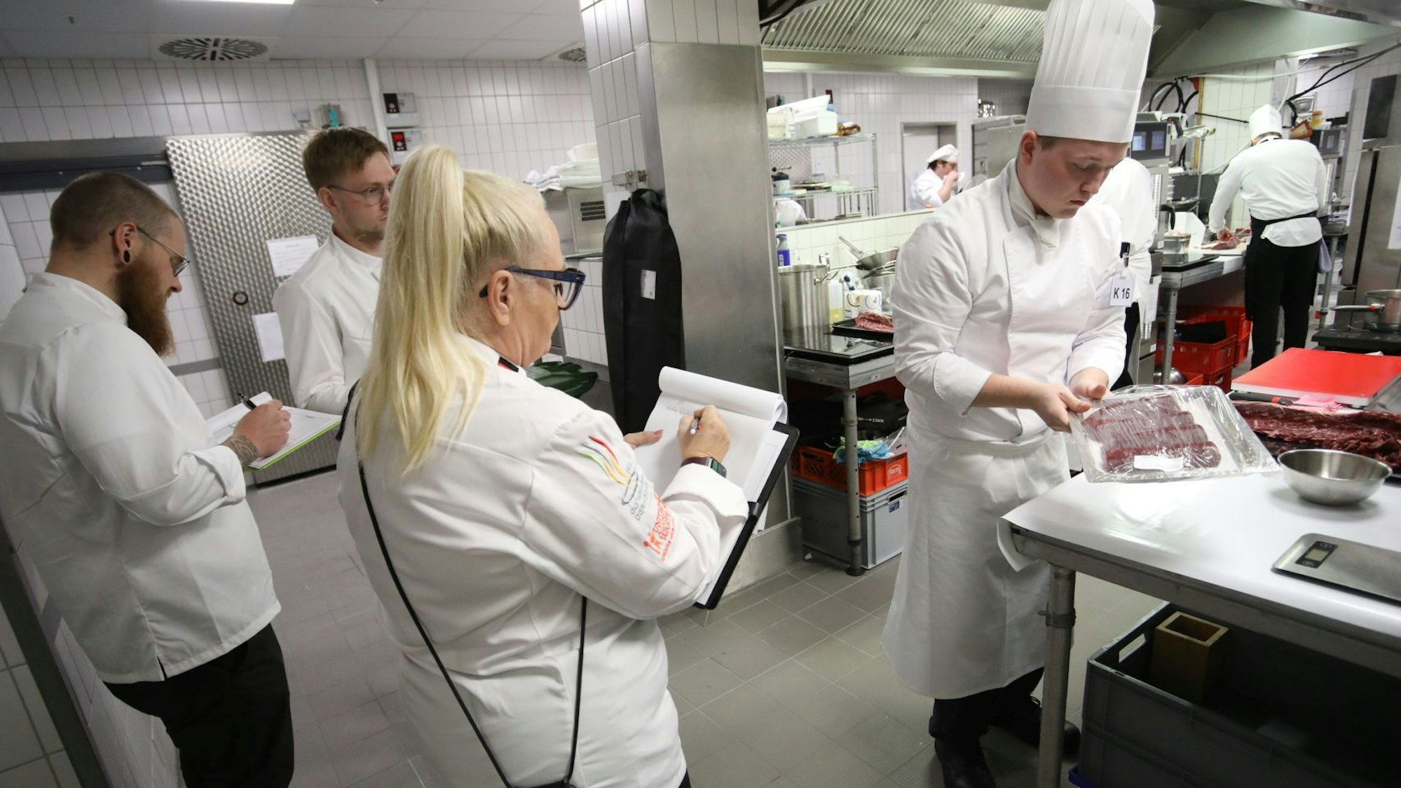 Blick in eine Hotelküche, drei Prüfer machen sich Notizen, während ein junger Koch ein Essen zubereitet.