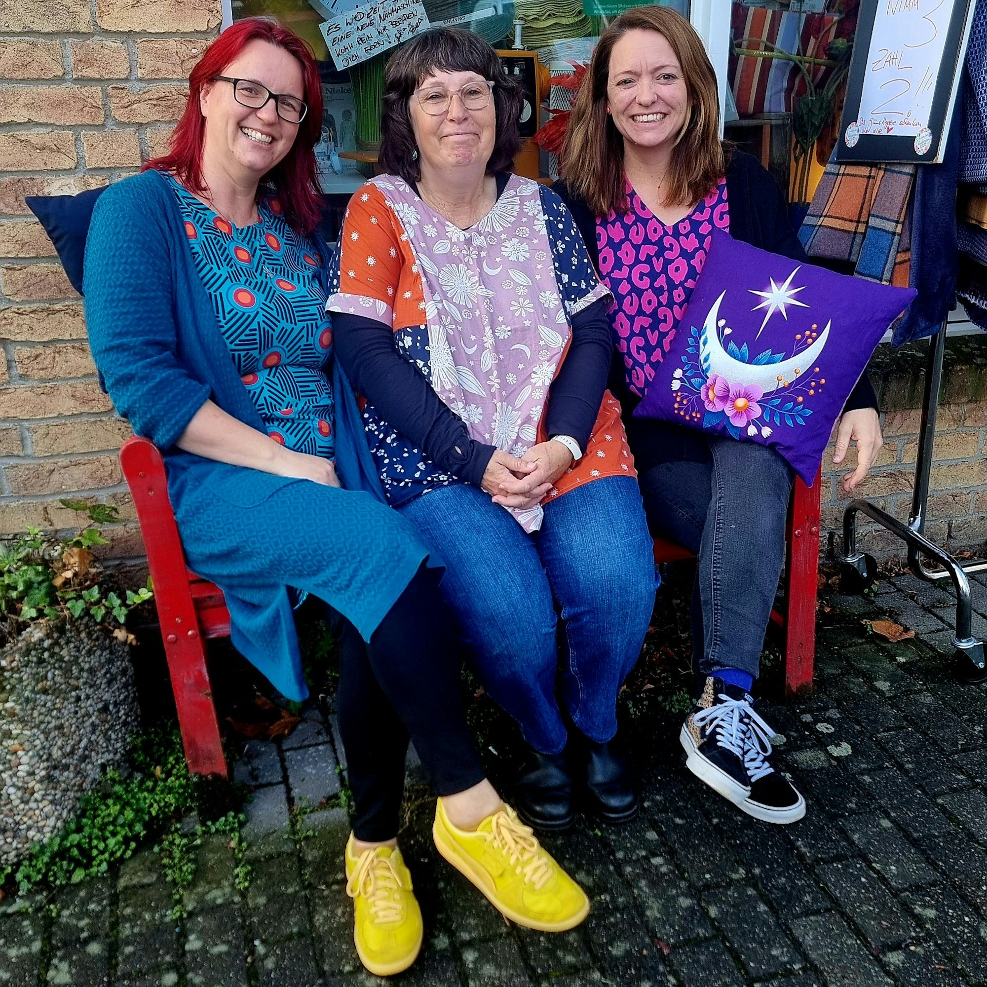 Drei Frauen sitzen auf einer roten Bank vor einem Stoffladen.