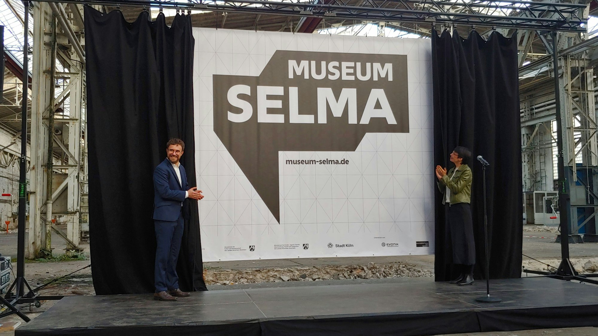 Zu sehen sind NRW-Integrationsministerin Josefine Paul (rechts) und Domid-Geschäftsführer Robert Fuchs (links), die auf einer Bühne stehen. Ein Vorhang hinter der Bühne ist geöffnet und zu sehen ist ein großes Plakat mit dem Schriftzug „Museum Selma“.