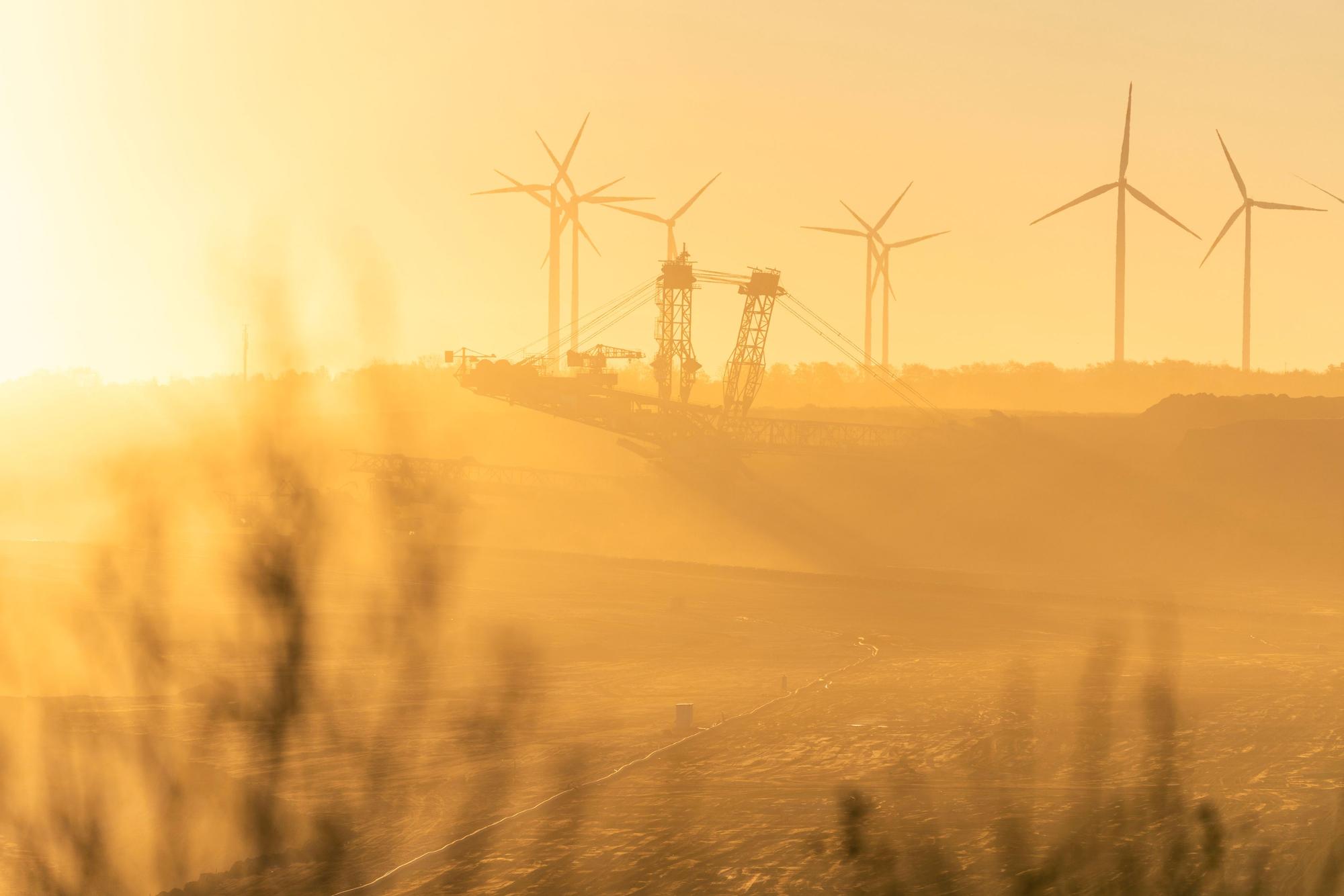 Das Bild ist durch den Sonnenaufgang tief orange, im Vordergrund ein paar verschwommene Sträucher, im Tagebau ein großer Bagger, und am Horizont einige Windräder.