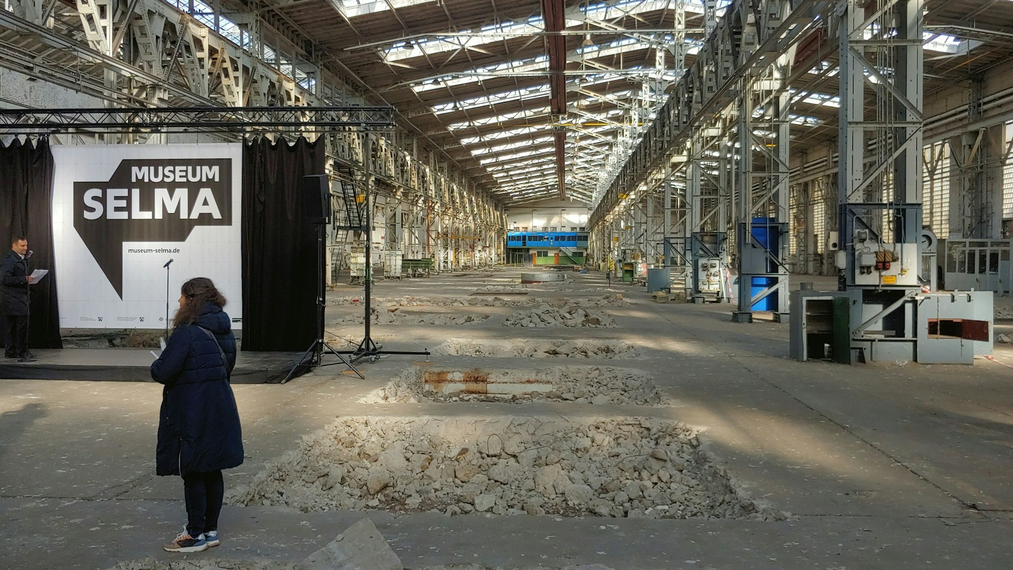 Zu sehen ist das Innere einer Halle der „Hallen Kalk“. Der Boden ist an mehreren Stellen aufgebrochen. Auf der linken Seite sind zwei Personen und eine Bühne zu erkennen.
