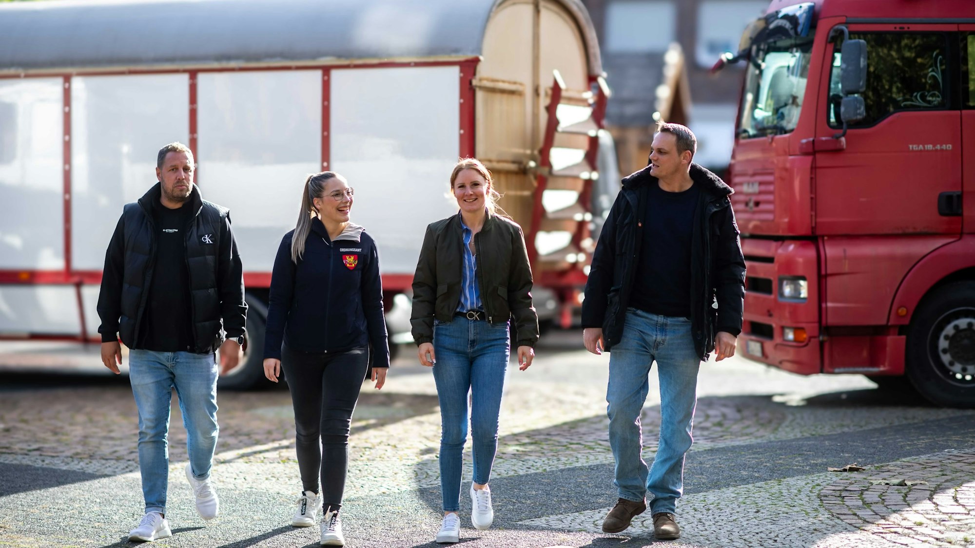 Zwei Männer und zwei Frauen der Euskirchener Ordnungsbehörde gehen über einen Platz. Hinter ihnen sind ein roter Lastwagen und eine rot-weiße Kirmesbude zu sehen.