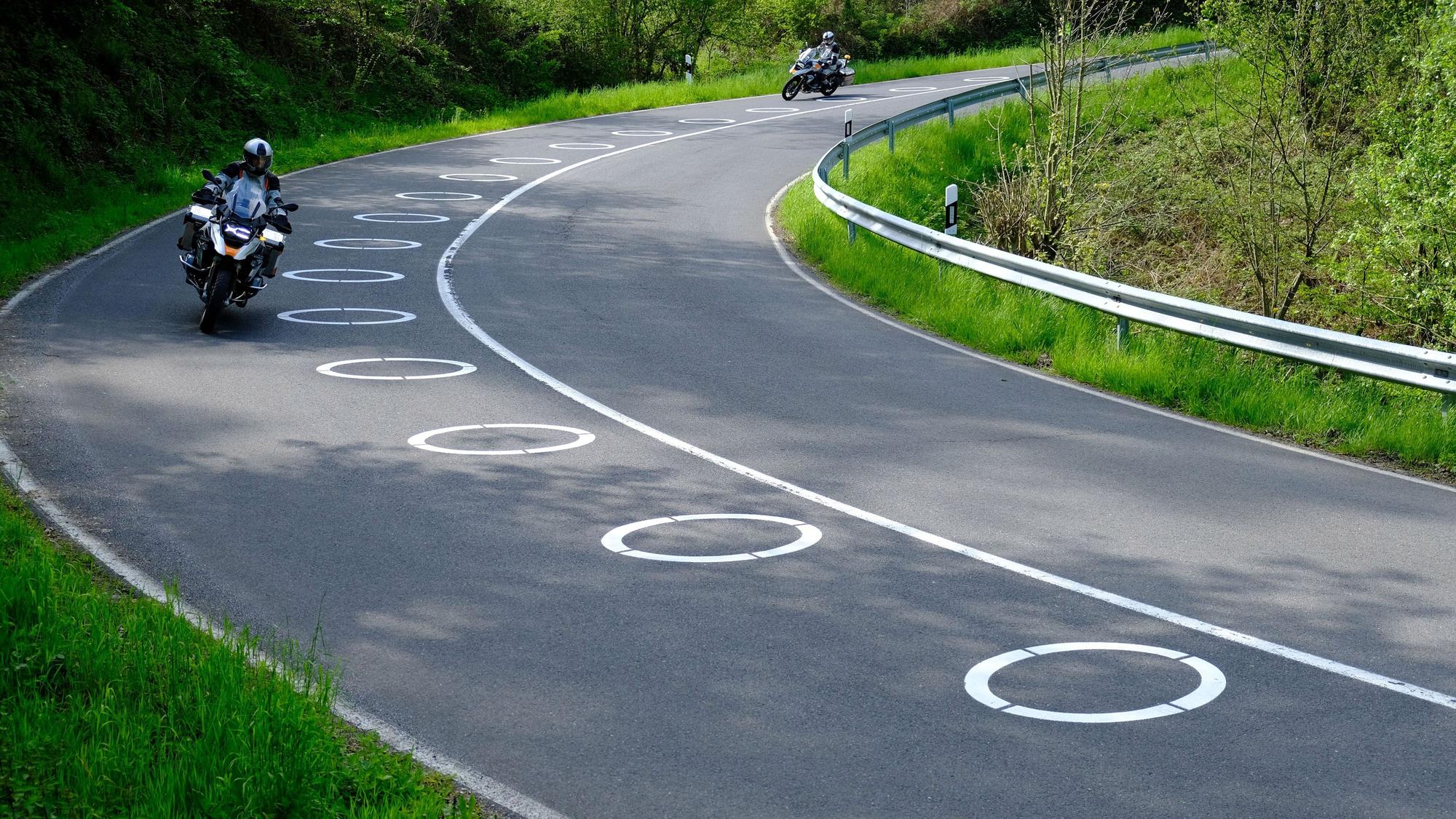 Auf einer Straße in der Eifel sind neben der Mittellinie ellipsenförmige Markierungen zu sehen. Zwei Motorradfahrer fahren daran vorbei.