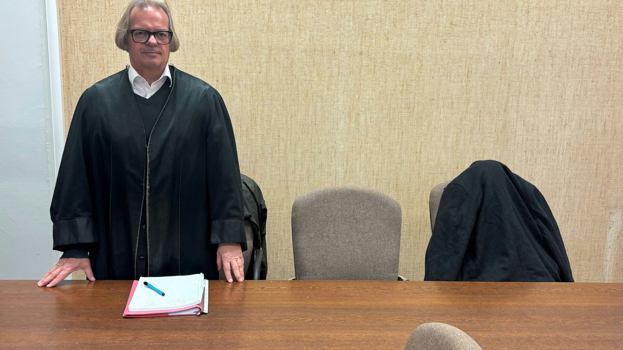 Der Verteidiger steht am Tisch, der Angeklagte sitzt auf einem Stuhl, verborgen unter einer Jacke.