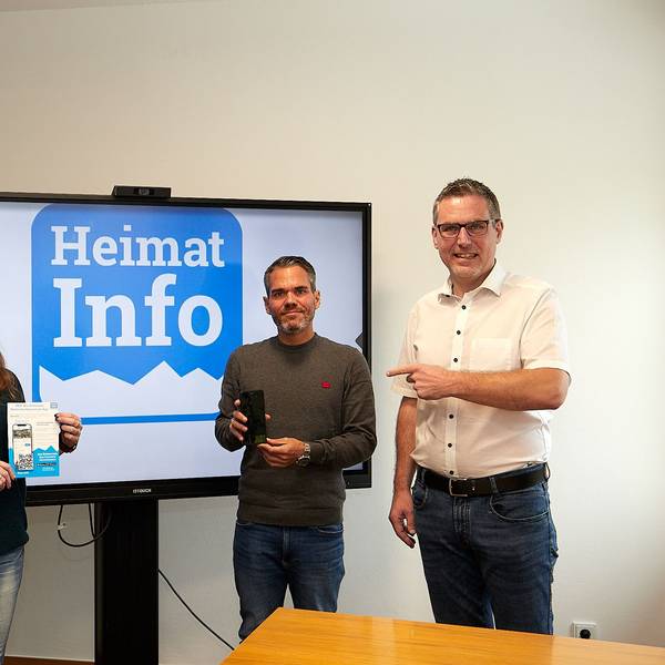 Zwei Frauen und zwei Männer stehen rechts und links von einem großen Bildschirm, auf dem in Blau auf weißem Grund das Symbol der App „Heimat Info“ zu sehen ist.