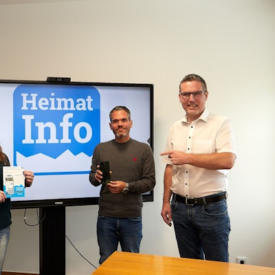 Zwei Frauen und zwei Männer stehen rechts und links von einem großen Bildschirm, auf dem in Blau auf weißem Grund das Symbol der App „Heimat Info“ zu sehen ist.