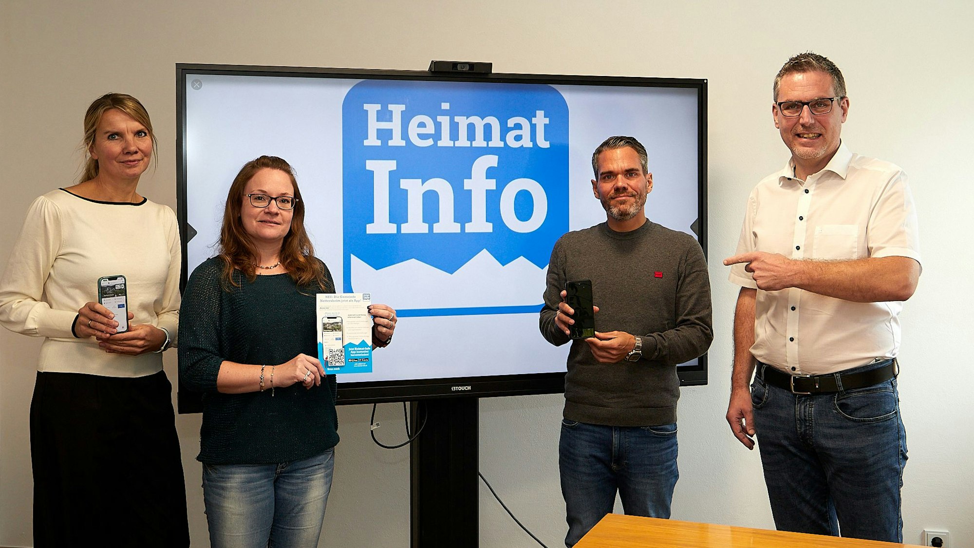 Zwei Frauen und zwei Männer stehen rechts und links von einem großen Bildschirm, auf dem in Blau auf weißem Grund das Symbol der App „Heimat Info“ zu sehen ist.