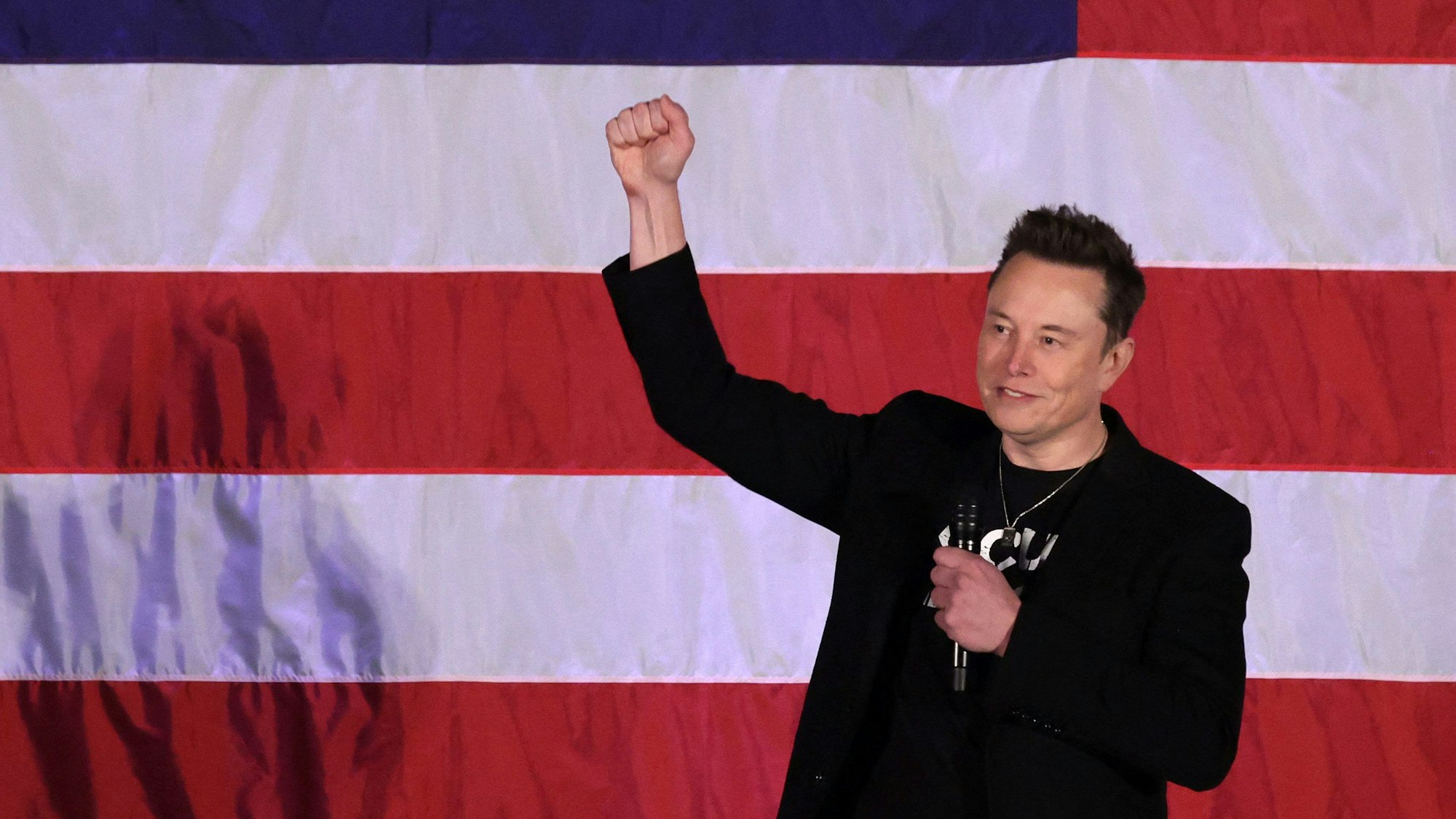 Elon Musk steht auf einer Wahlkampfveranstaltung mit erhobener Faust vor einer US-Flagge.