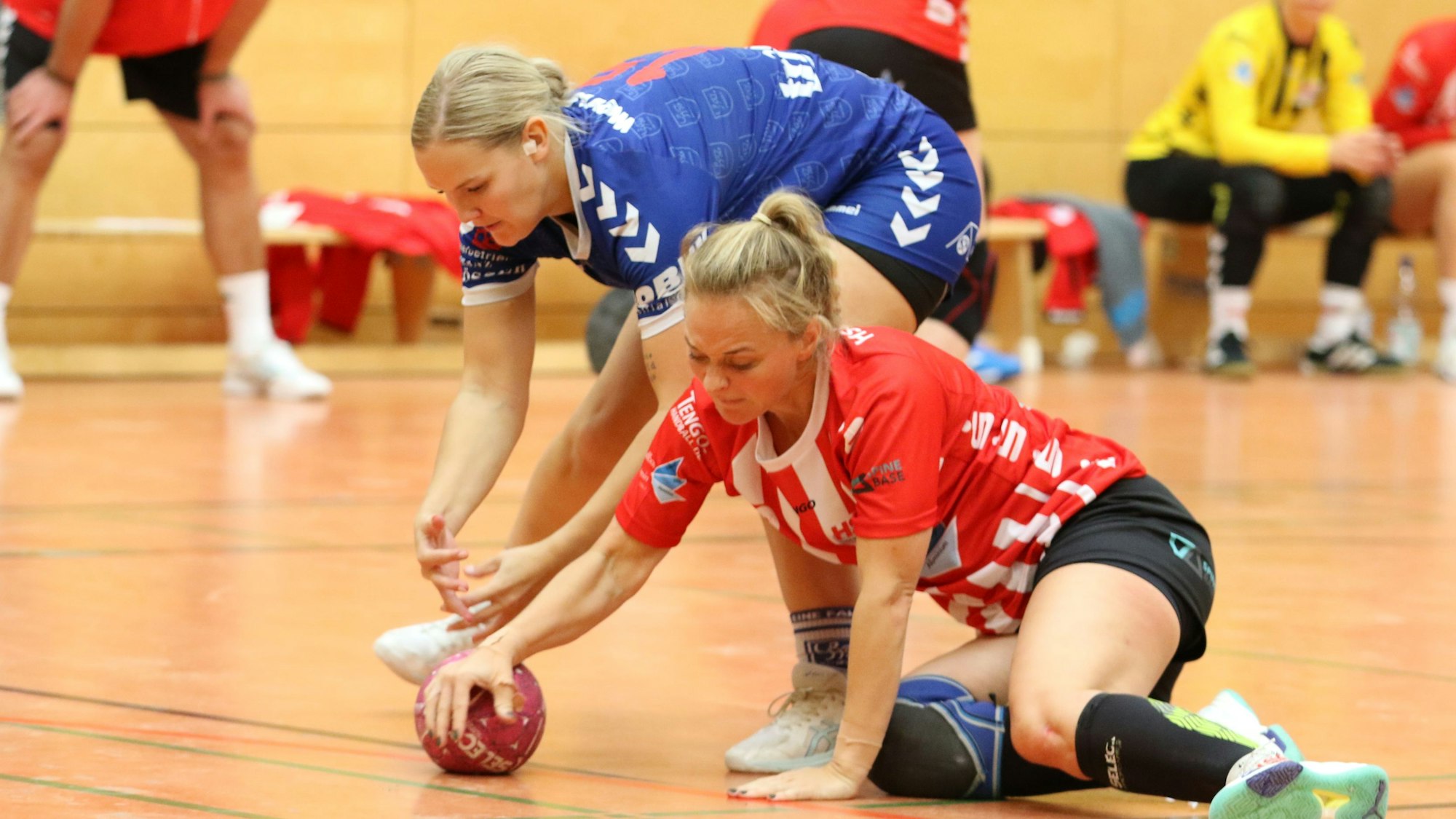 Eine Handballerin, die auf dem Boden hockt, sichert einen Ball vor einer Gegenspielerin.
