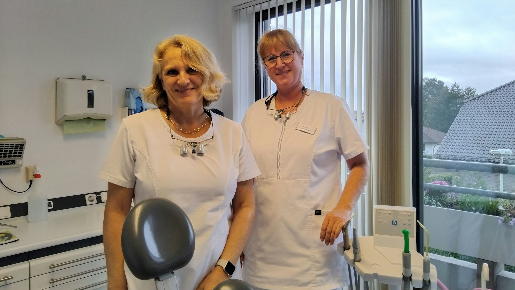 Zwei Zahnärztinnen in weißen Kitteln stehen in einem Behandlungszimmer.