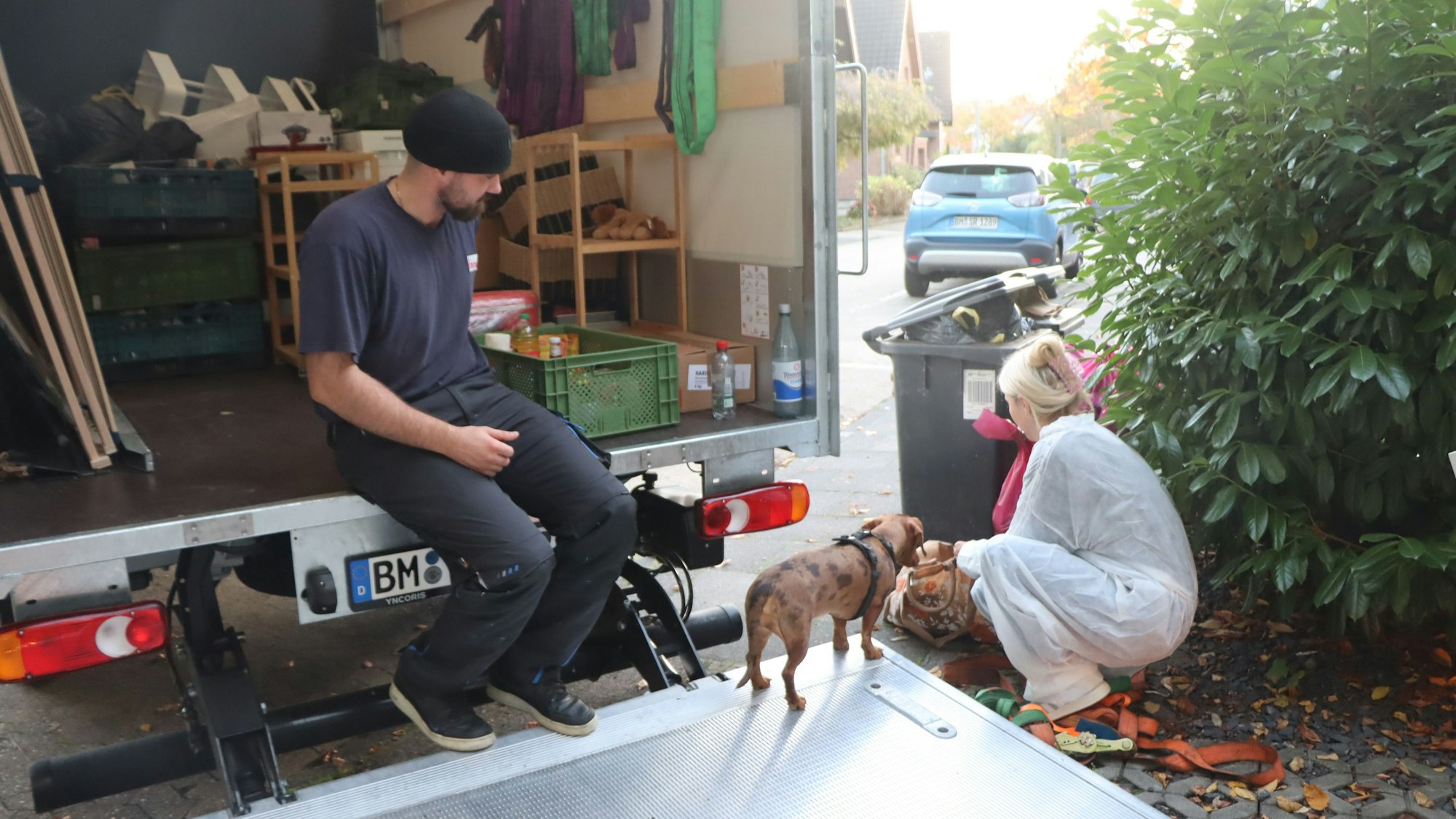 Ein Mann sitzt auf der Ladefläche eines Lkw, neben der Rampe hockt eine Frau im Schutzanzug neben einem Hund.