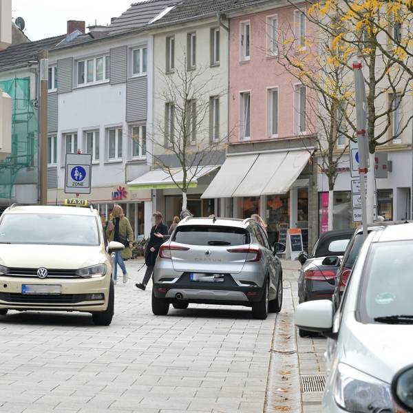 Am Straßenrand parken Autos. In der Mitte der Fußgängerzone begegnen sich zwei Autos.