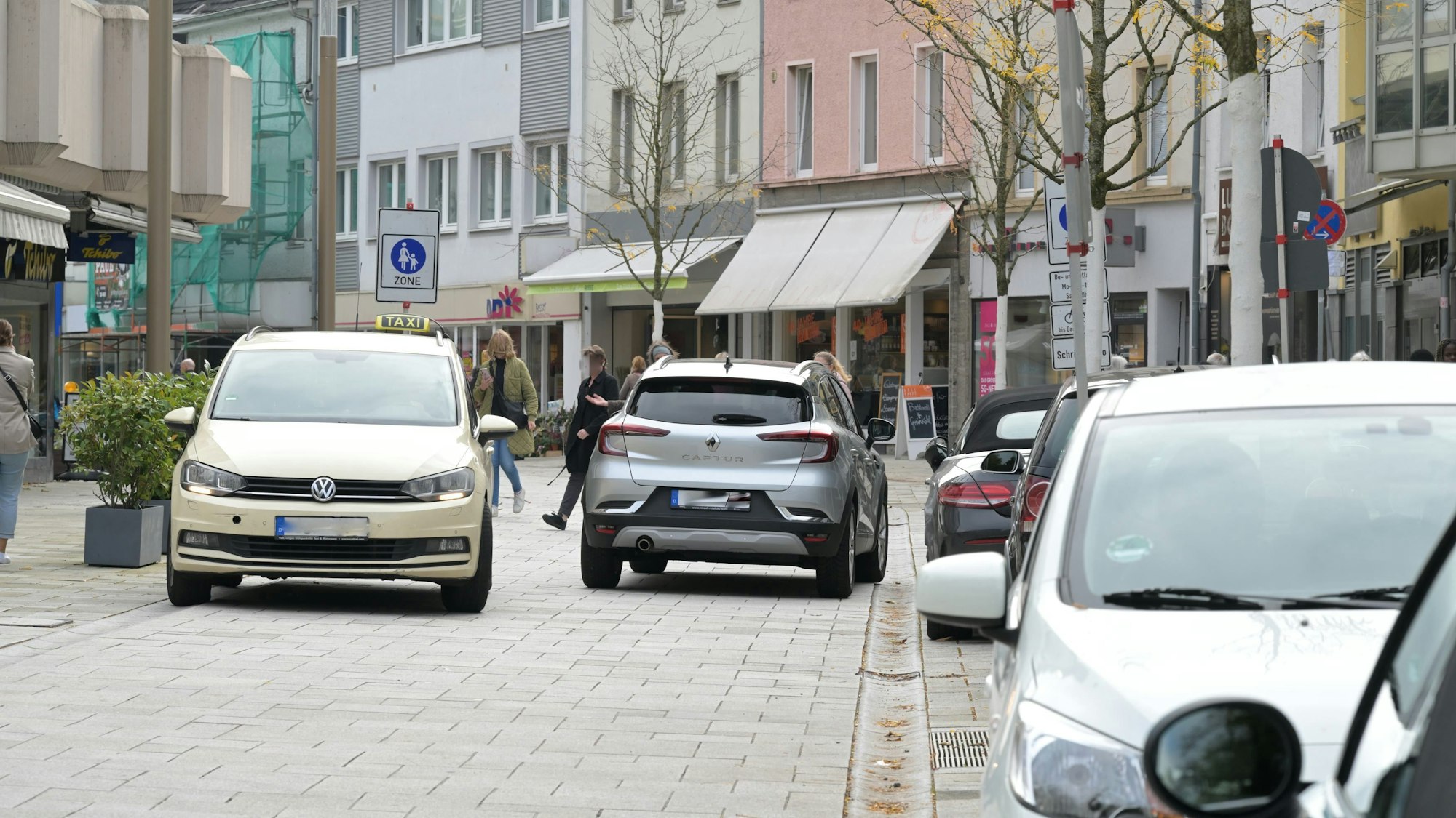 Am Straßenrand parken Autos. In der Mitte der Fußgängerzone begegnen sich zwei Autos.