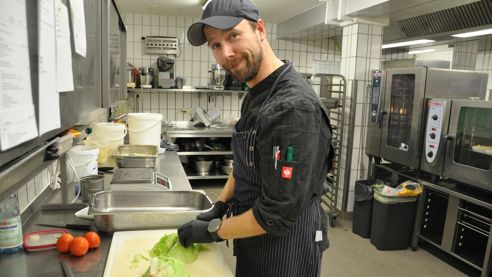 Der Koch trägt schwarze Kleidung, eine Mütze und Handschuhe, während er in der Kantine Salat schnippelt.