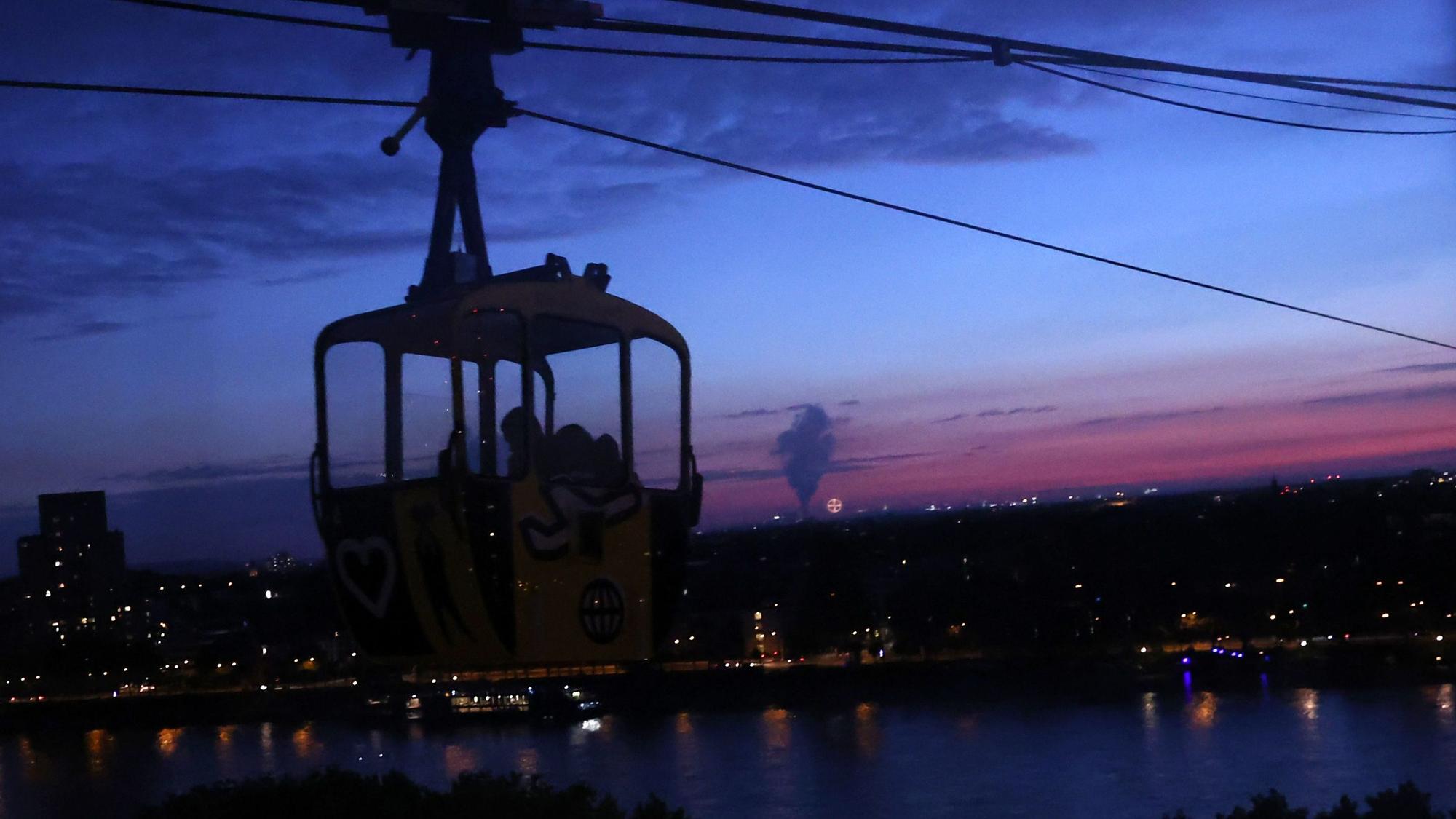 Kölner Seilbahn fährt bei Nacht. Im Hintergrund ist der Rhein zu sehen.