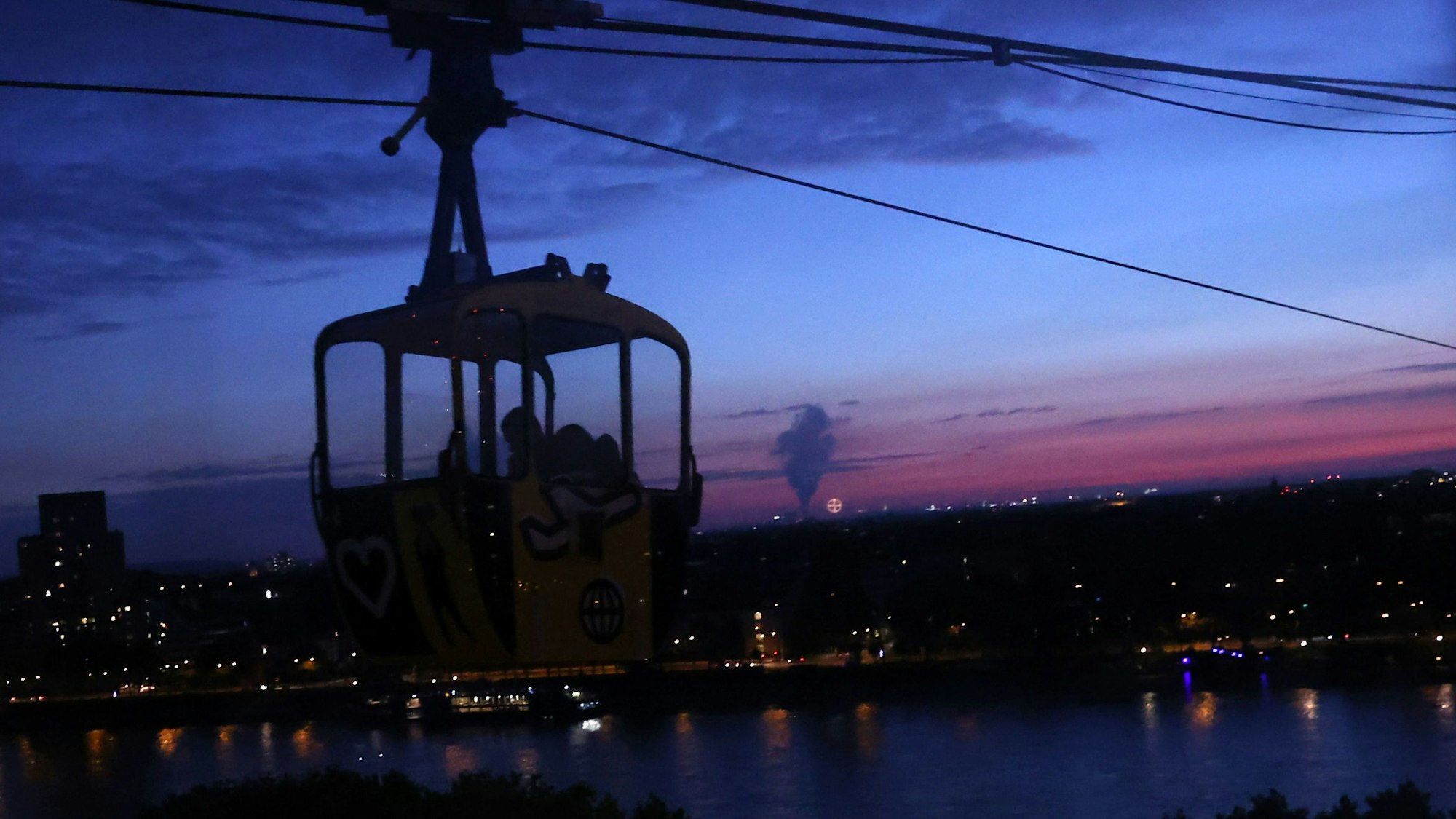 Kölner Seilbahn fährt bei Nacht. Im Hintergrund ist der Rhein zu sehen.