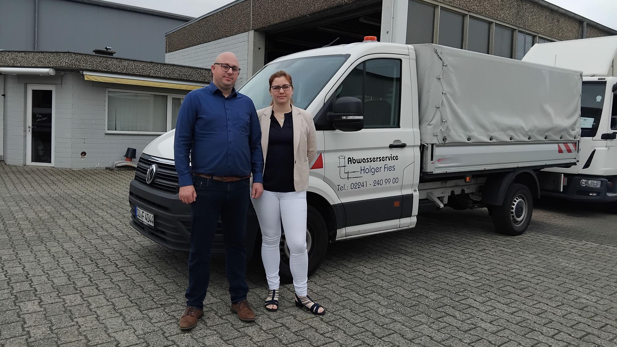 Ein Mann und eine Frau stehen vor einem Wagen mit Anhänger, auf dem "Abwasserservice Holger Fies" steht.