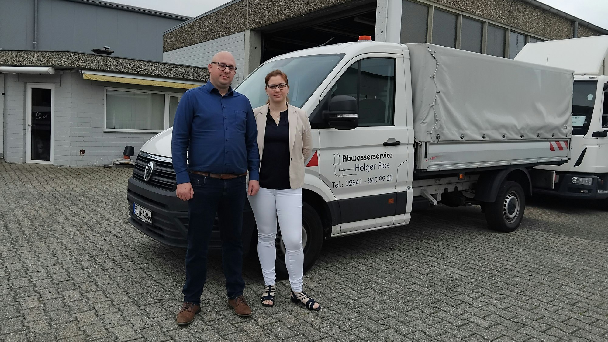 Ein Mann und eine Frau stehen vor einem Wagen mit Anhänger, auf dem "Abwasserservice Holger Fies" steht.