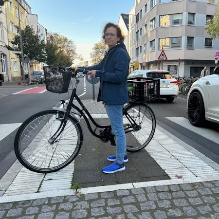Anwohnerin Monika Bodenstein steht im Oktober 2024 mit Fahrrad auf der Verkehrsinsel Vogelsanger Straße/Sömmeringstraße in Ehrenfeld