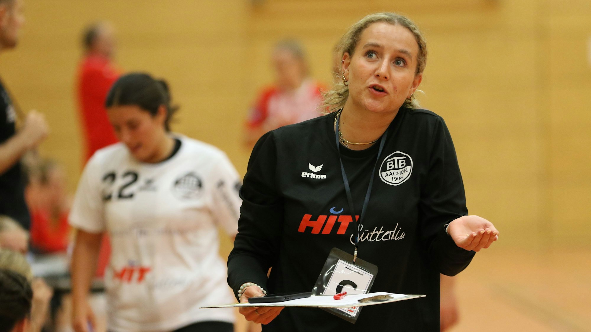 Die Betreuerin von BTB Aachen hält die linke Hand zur Mulde. Nicht zu sehen ist, dass dort ein Zahn liegt. Auf dem Klemmbrett liegt ein blutiger Wattebausch. Die Betreuerin blickt zur Tribüne hoch.