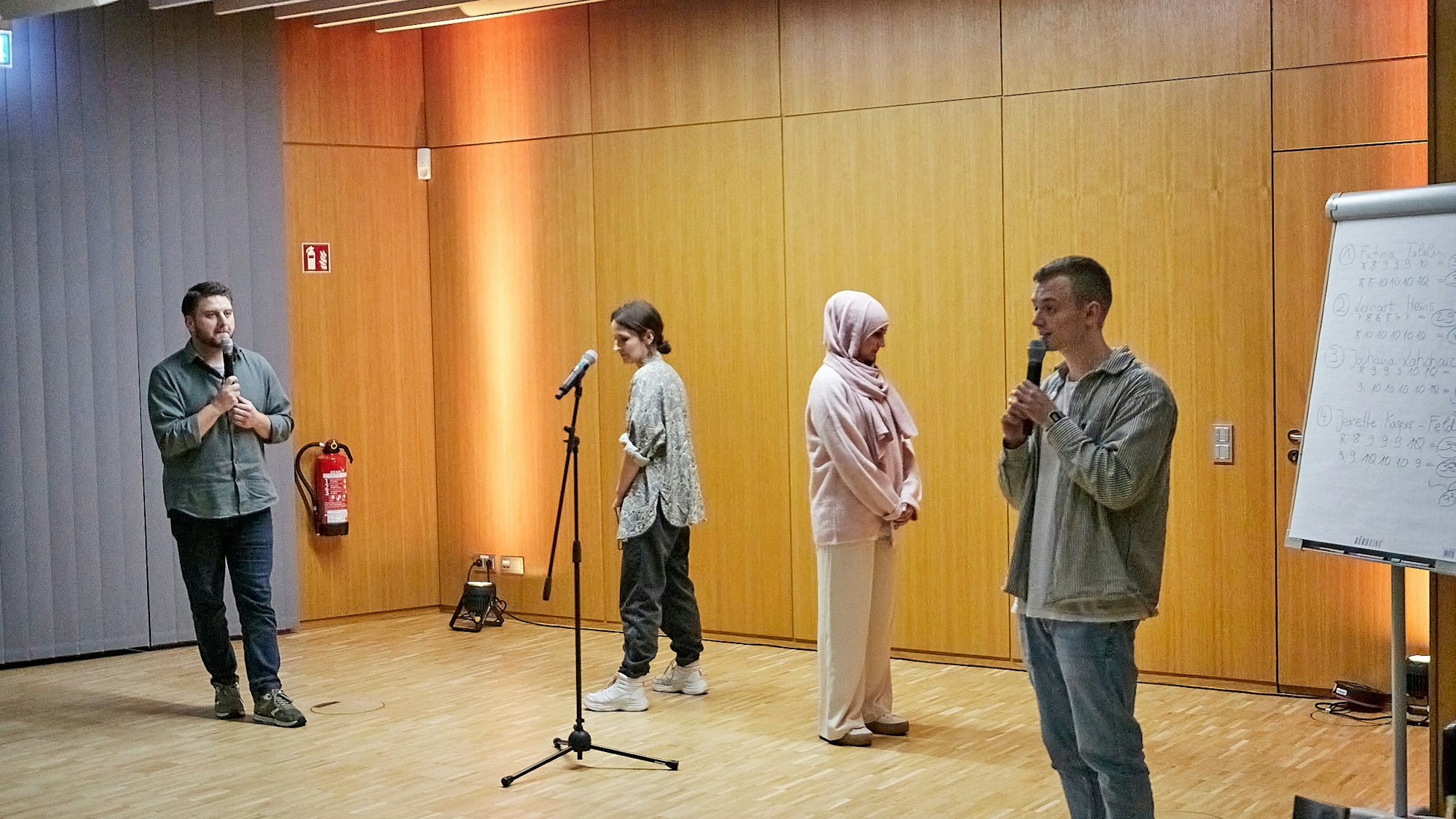 Zwei Frauen, die einander den Rücken zuwenden, und zwei Männer mit Mikrofonen stehen in einem Raum.
