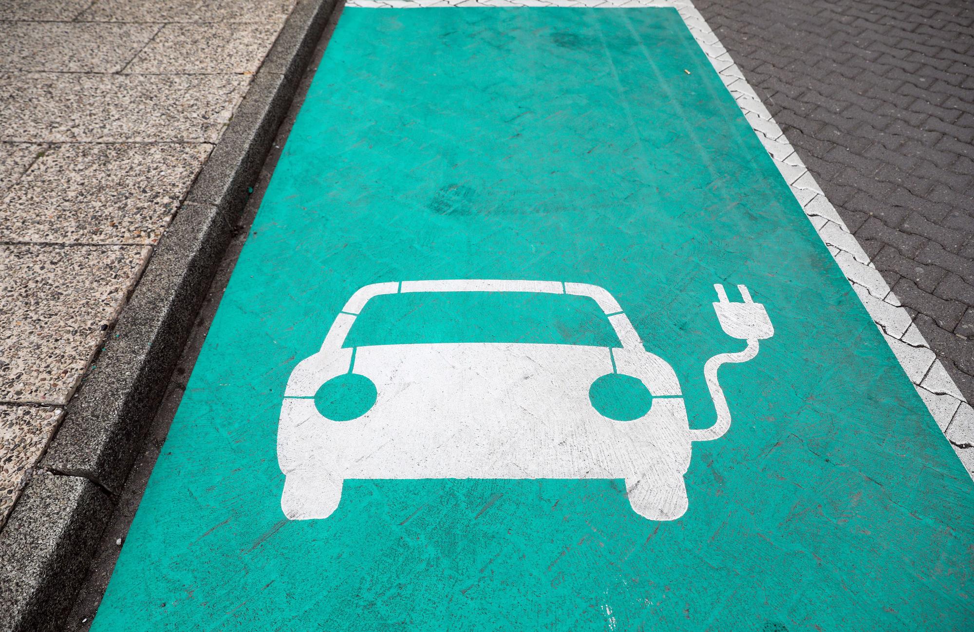 Ein stilisiertes Auto auf einem grünen Grund markiert einen Parkplatz mit Ladesäule für Elektrofahrzeuge.