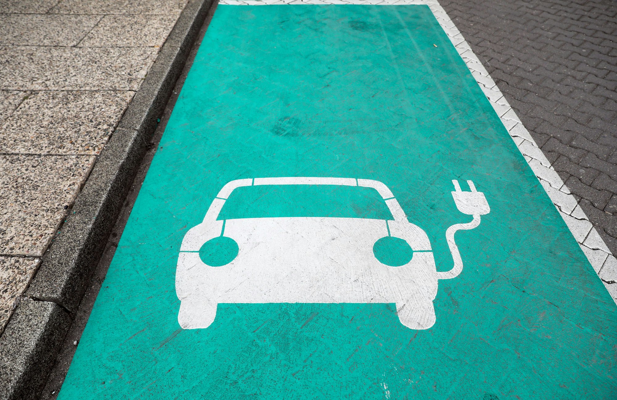 Ein stilisiertes Auto auf einem grünen Grund markiert einen Parkplatz mit Ladesäule für Elektrofahrzeuge.