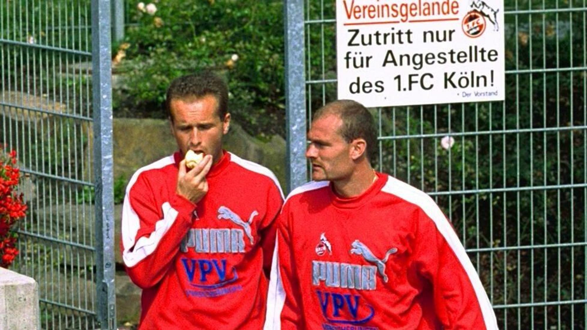 Dirk Lottner beißt in einen Apfel. Ralf Hauptmann läuft neben ihm aus dem FC-Gelände. Die beiden tragen rote Vereinspullis.