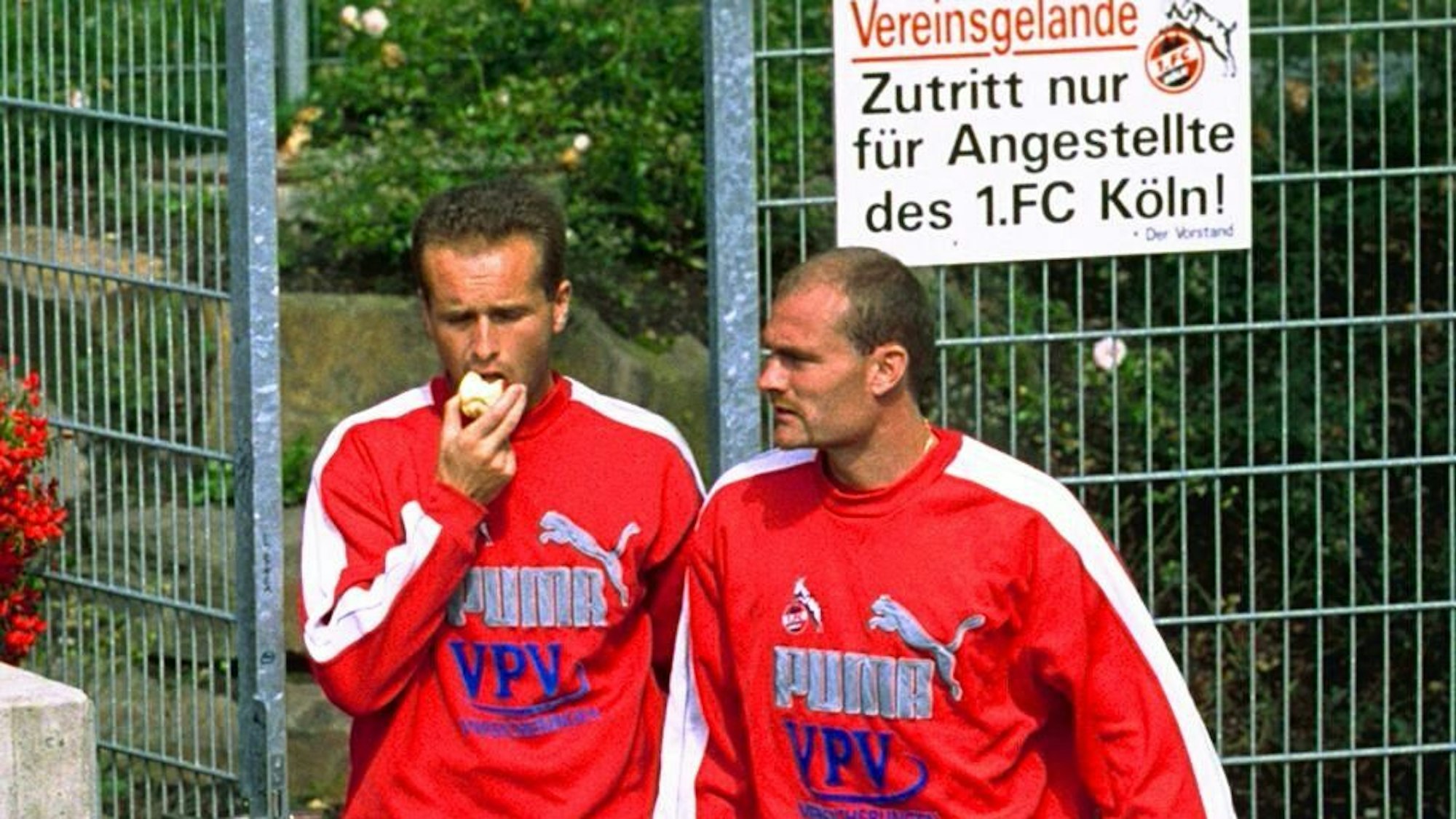 Dirk Lottner beißt in einen Apfel. Ralf Hauptmann läuft neben ihm aus dem FC-Gelände. Die beiden tragen rote Vereinspullis.