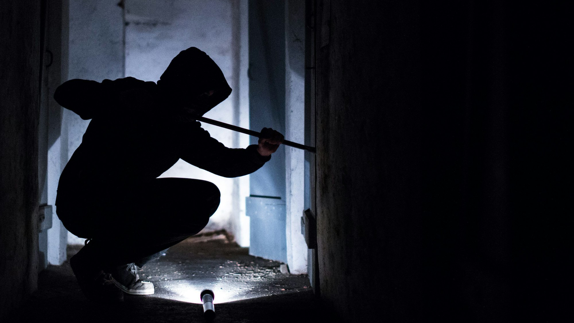 Eine Person mit einer Kapuze über dem Kopf steigt mit einem Brecheisen in ein Fenster ein.