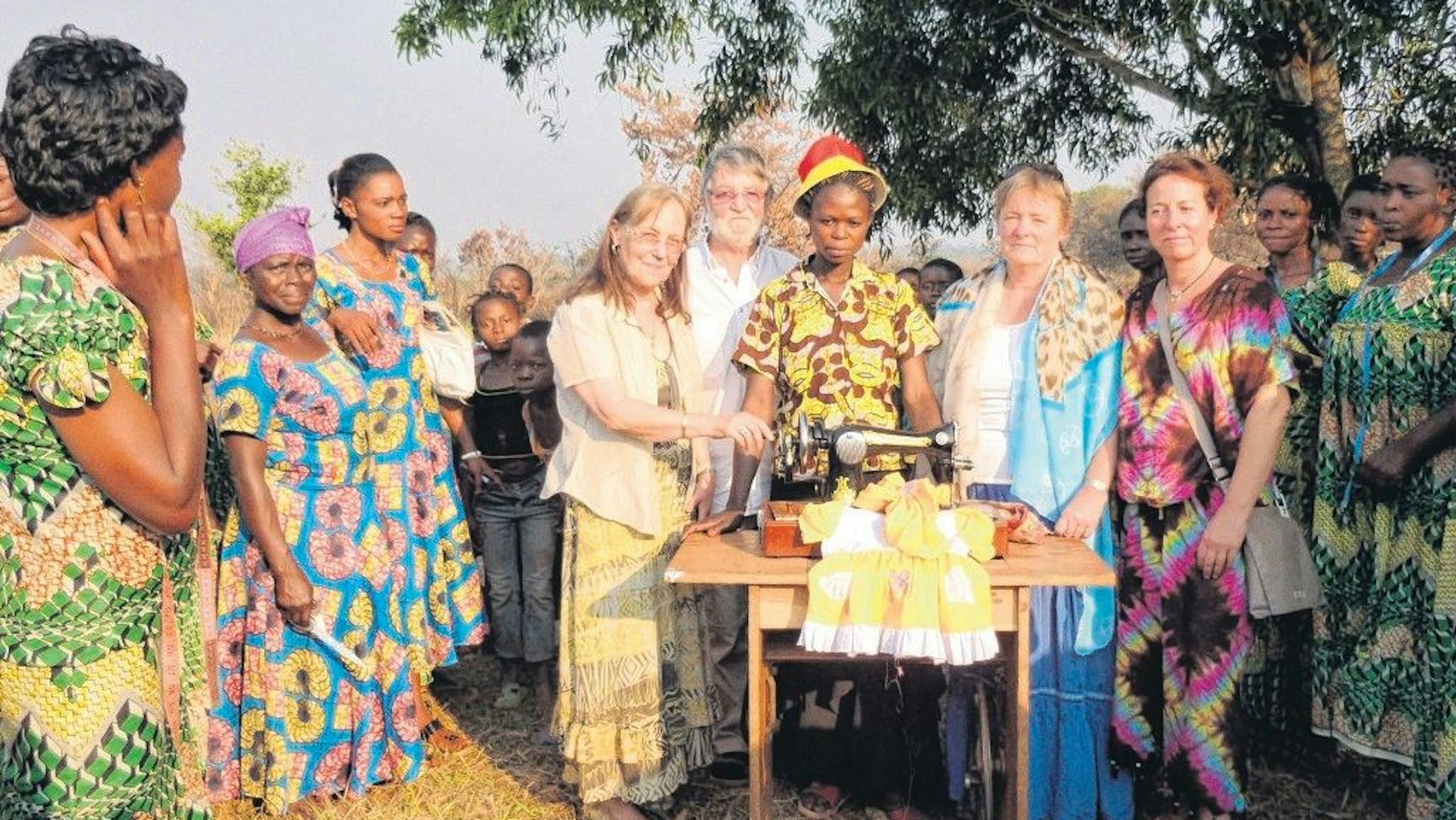 Vier Besucher aus Hennef stehen in einem Kreis afrikanischer Frauen, die bunte Gewänder tragen. In der Mitte steht eine Afrikanerin an einer alten Nähmaschine.