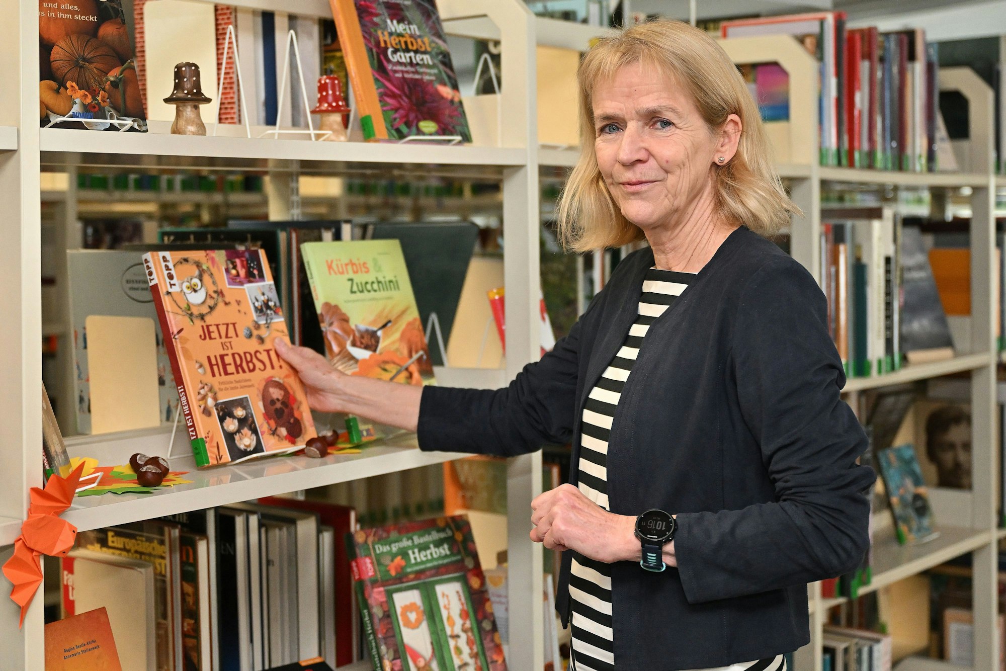 Die Büchereileiterin steht vor einem Bücherregal und hält ein Buch zum Thema Herbst in der Hand.