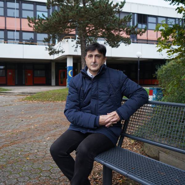 Das Foto zeigt einen Mann, der auf einer Bank vor einem Schulblock sitzt.