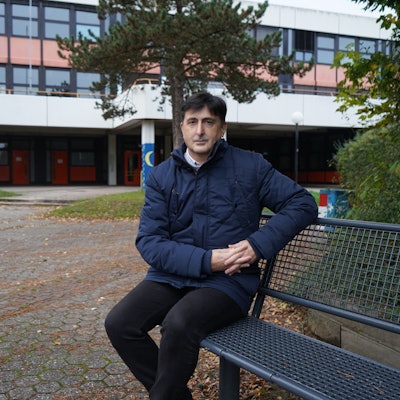 Das Foto zeigt einen Mann, der auf einer Bank vor einem Schulblock sitzt.