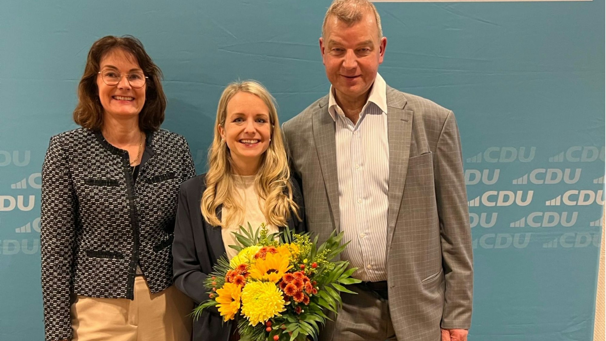 CDU-Bürgermeisterkandidatin Laura Lundberg steht mit der CDU-Fraktionsvorsitzenden im Odenthaler Gemeinderat, Nicola Ciliax-Kindling, und Odenthals CDU-Parteivorsitzendem Christof Jochum vor einer blauen CDU-Wand.