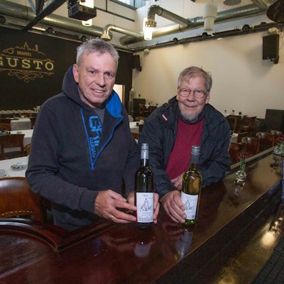Zwei Männer stehen am Tresen einer Gaststätte und halten zwei Flaschen mit Wein in Händen.