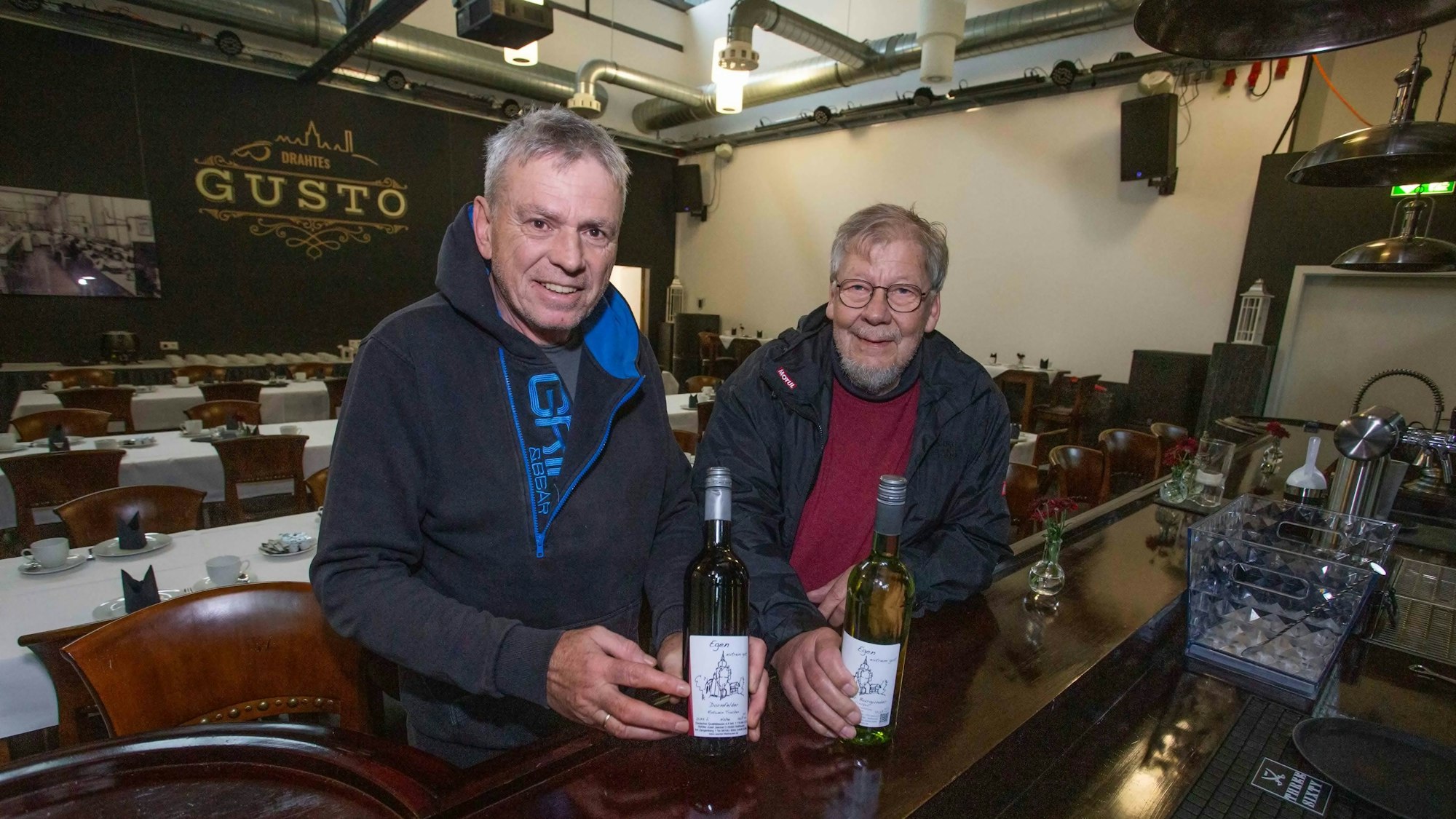 Zwei Männer stehen am Tresen einer Gaststätte und halten zwei Flaschen mit Wein in Händen.
