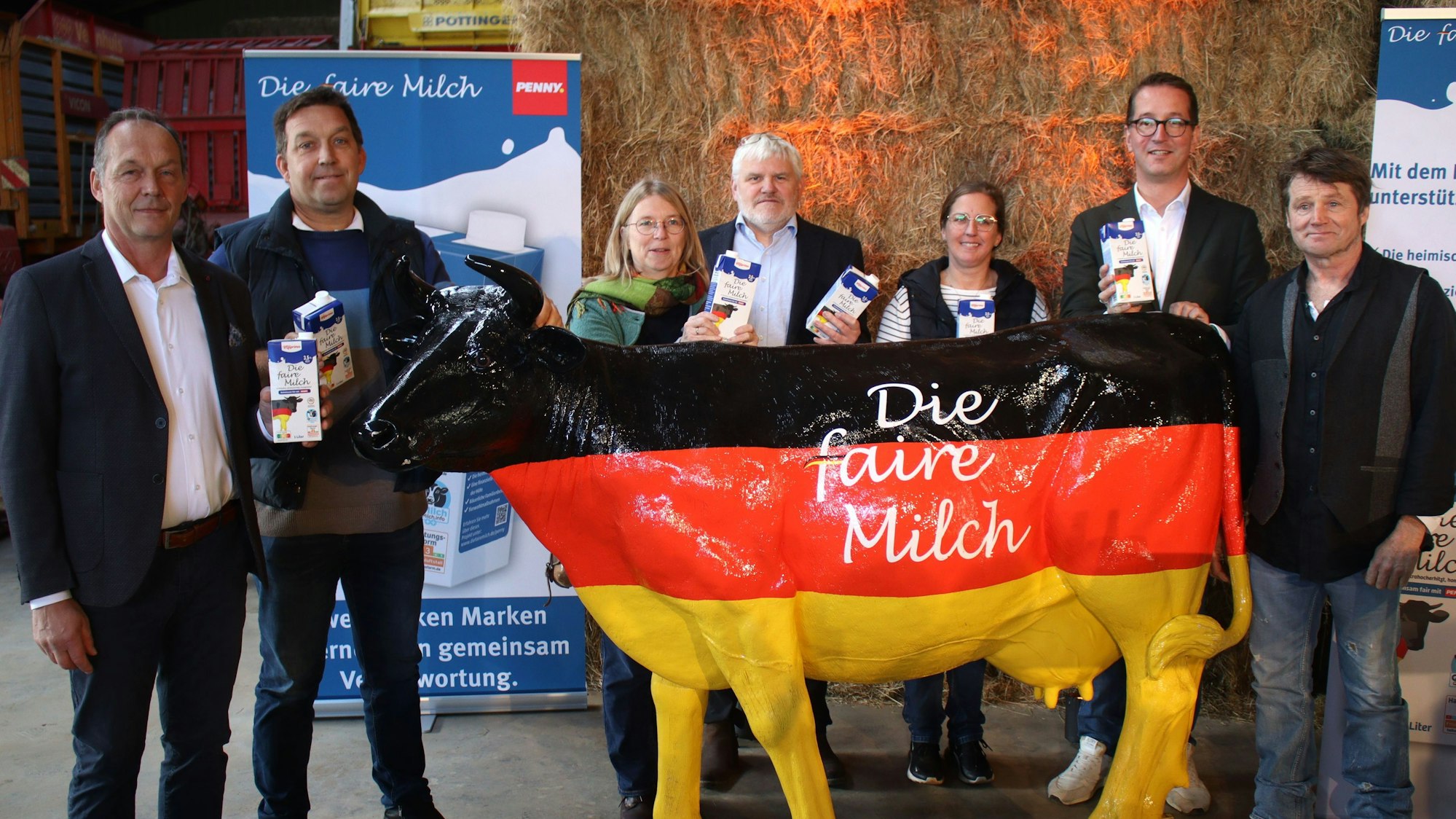 Die Vertragsunterzeichner stehen vor Strohballen und hinter einer schwarz-rot-goldenen Kuh. Einige halten Milchtüten in der Hand.