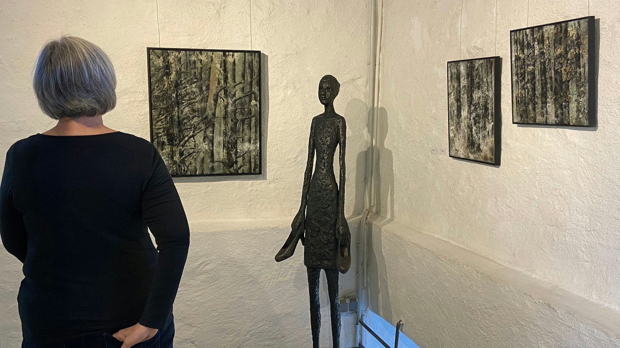 Zu sehen ist eine Frau, die auf Bilder an einer Wand schaut. Vor der Wand steht auch eine weibliche Skulptur.