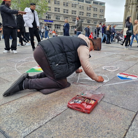 Ein Straßenmaler auf der Domplatte zeichnet mit einer Schablone Herzen auf und malt sie in der Farbe von verschiedenen Länderflaggen aus. (Archivbild)