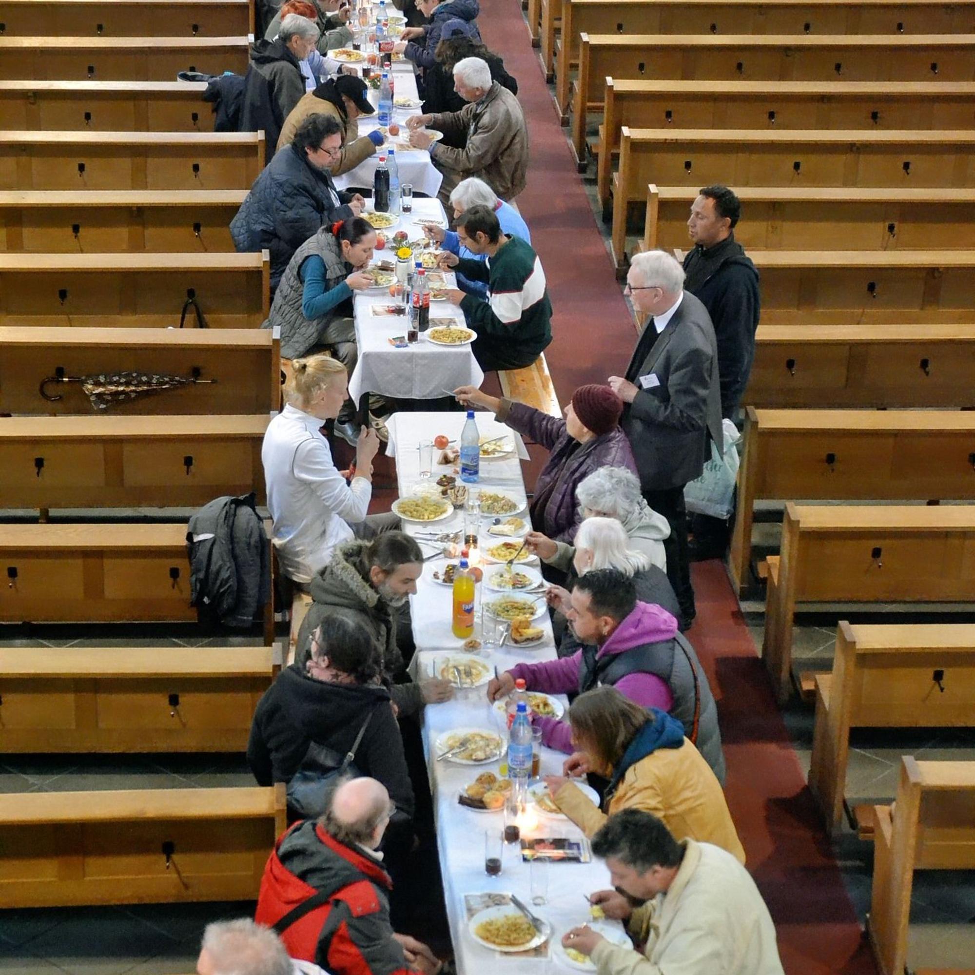 Zahlreiche Männer und Frauen sitzen an einer langen Tafel und essen. Anhand der Bänke rechts und links von ihnen ist zu erkennen, dass sie in einer Kirche sitzen.