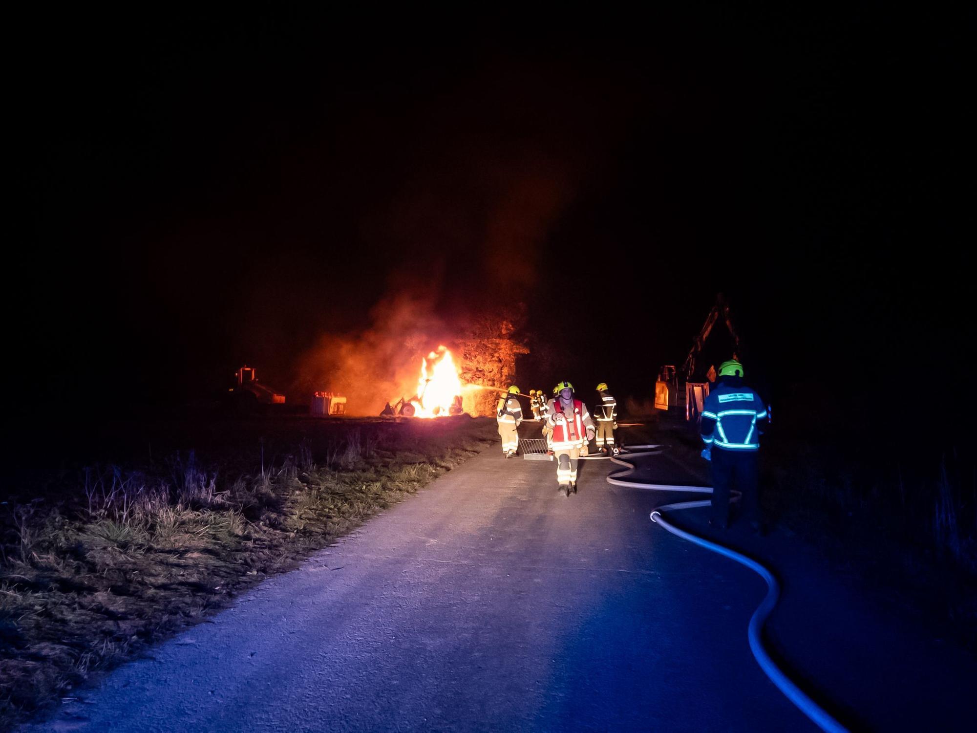 Ein Feuer im Dunkeln, im Vordergrund mehrere Einsatzkräfte und ein Feuerwehrschlauch.